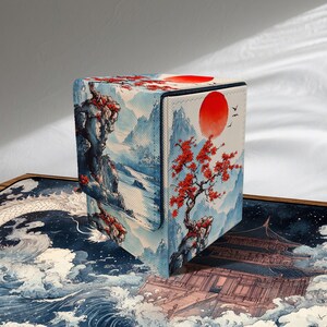 Puede incluir: Un estuche cuadrado para tarjetas con un diseño de paisaje japonés. El estuche presenta un sol rojo, un árbol con hojas rojas y una escena de montaña en tonos azules y blancos. El estuche tiene un cierre de solapa magnética.