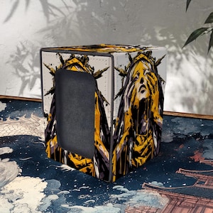 May include: A square, white box with a black, yellow, and gray graphic design. The design features a stylized face with a screaming expression. The box has a rectangular opening on one side and is placed on a patterned surface.