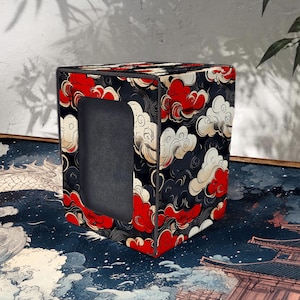 Peut inclure: Une boîte de rangement cubique avec un intérieur noir et un extérieur à motifs de nuages rouges, blancs et noirs. La boîte a une ouverture rectangulaire sur un côté. Le design rappelle l'art japonais.