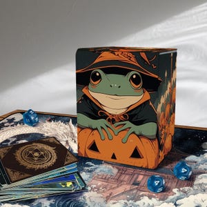 Puede incluir: Una caja decorativa con una rana de dibujos animados con un sombrero de bruja y una calabaza. La caja tiene un tema de Halloween y está rodeada de dados azules y una baraja de cartas.