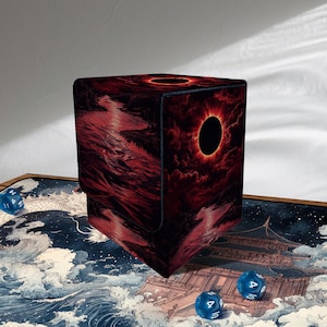May include: A black and red dice box featuring a dramatic illustration of a solar eclipse. The box is cube-shaped and has a dark, textured appearance. Several blue dice are scattered around the box, suggesting a tabletop gaming theme.