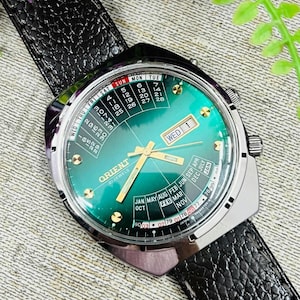 May include: A vintage Orient watch with a green dial and gold-tone hands and markers. The watch features a calendar function and is set to Wednesday, the 1st. It has a stainless steel case and a black leather strap.