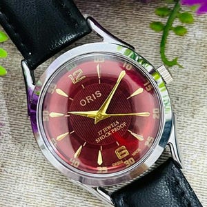 Może przedstawiać: Zegarek na rękę Oris w stylu vintage z bordową tarczą i złotymi wskazówkami. Zegarek ma kopertę ze stali nierdzewnej i czarny skórzany pasek. Tarcza zawiera napis "17 JEWELS SHOCK PROOF". Klasyczny zegarek.