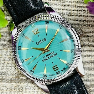 Peut inclure: Une montre Oris vintage avec un cadran turquoise, des aiguilles dorées et un bracelet en cuir noir. Le cadran affiche le texte "ORIS", "17 JEWELS" et "SHOCK PROOF". La montre a une lunette argentée et une couronne texturée.