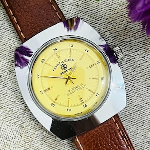 Peut inclure: Une montre vintage Favre-Leuba Geneve avec un cadran doré et un bracelet en cuir marron. La montre a une lunette carrée argentée avec des accents violets. Le cadran affiche le texte "17 JEWELS SHOCK PROOF".