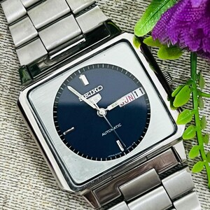 Könnte beinhalten: Eine silberfarbene Seiko Automatik-Armbanduhr mit quadratischem Zifferblatt und dunkelblauem Zifferblatt. Die Uhr hat ein silbernes Armband und zeigt Tag und Datum an. Die Uhr ist auf Sonntag, den 11. eingestellt.