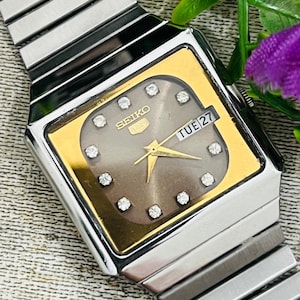 Könnte beinhalten: Eine Vintage Seiko-Uhr mit silbernem Armband und einem goldfarbenen quadratischen Zifferblatt. Das Zifferblatt ist grau mit diamantartigen Markierungen und goldenen Zeigern. Der Tag wird als "TUE 27" angezeigt.