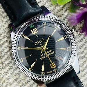Könnte beinhalten: Eine Vintage-Armbanduhr von Oris mit schwarzem Zifferblatt und goldfarbenen Zeigern und Markierungen. Die Uhr hat eine silberfarbene Lünette und ein schwarzes Lederarmband. Auf dem Zifferblatt steht "ORIS", "17 JEWELS" und "SHOCK PROOF".
