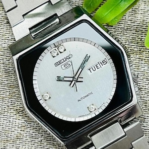 Könnte beinhalten: Eine silberne Seiko 5 Automatik-Uhr mit quadratischem Zifferblatt und silbernem Armband. Das Zifferblatt ist hellgrau mit Diamant-Stundenmarkierungen und Wochentag und Datum. Die Uhr zeigt Dienstag, den 15.