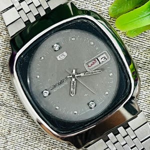 Könnte beinhalten: Eine silberfarbene Armbanduhr mit grauem Zifferblatt und Edelstahlarmband. Das Zifferblatt der Uhr hat rautenförmige Stundenmarkierungen und eine Datumsanzeige. Die Uhr ist mit "SEIKO AUTOMATIC" beschriftet.