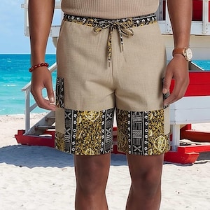 Puede incluir: Pantalones cortos de color beige con cintura de cordón y paneles laterales con un diseño estampado en negro y dorado. Los pantalones tienen una sección central de color beige liso y un borde estampado a juego en la cintura, sobre la rodilla.