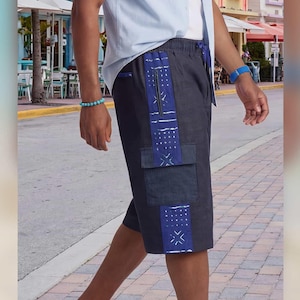 May include: Dark charcoal gray shorts featuring a vertical side panel with a blue and white geometric pattern, a zippered pocket, and a large cargo pocket. The shorts have an elastic waistband with a drawstring and are paired with black sandals.