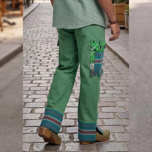 May include: Green pants with patterned patches and striped cuffs. The pants have a relaxed fit and are made of a durable fabric. The cuffs feature teal, red, and white stripes. The pants are paired with brown clogs.