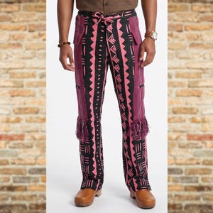 May include: Black and pink patterned pants with a drawstring waist and fringe detail at the hem. The pants feature a geometric design and vertical stripes. The model is wearing brown clogs.