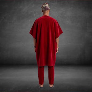 May include: A red velvet traditional African outfit, including a tunic and pants. The outfit is worn with a patterned head wrap and beaded necklace. The person is viewed from the back, set against a dark gray background.