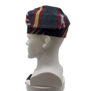 May include: A flat cap with a striped pattern in red, navy blue, yellow, and white. The cap is displayed on a white mannequin head. The cap has a short brim and a flat top.