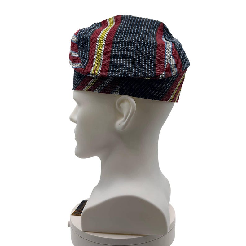 May include: A flat cap with a striped pattern in red, navy blue, yellow, and white. The cap is displayed on a white mannequin head. The cap has a short brim and a flat top.