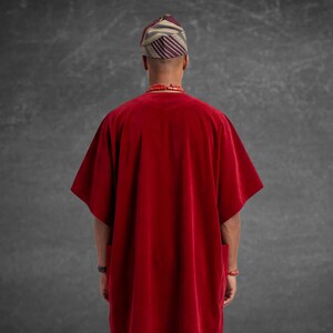 May include: A person wearing a red velvet tunic with wide sleeves, a striped head wrap, and a red beaded necklace. The back view shows the tunic's simple design and the traditional headwear.