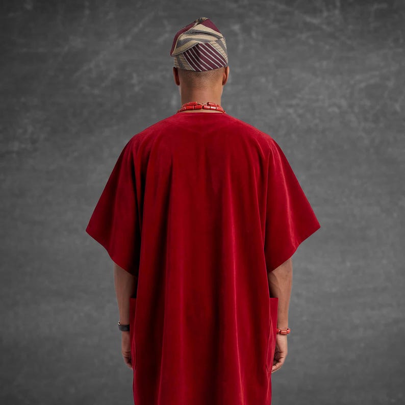 May include: A person wearing a red velvet tunic with wide sleeves, a striped head wrap, and a red beaded necklace. The back view shows the tunic's simple design and the traditional headwear.