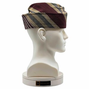May include: A patterned hat with burgundy and gold stripes, displayed on a white mannequin head. The hat features a rectangular crown and a brim that wraps around the head. The stripes are diagonal and create a visually appealing design.