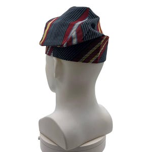 May include: A folded hat with a striped pattern in navy, red, and gold. The hat is displayed on a white mannequin head, highlighting the fabric's texture and design. The stripes on the hat run vertically, creating a striking visual effect.