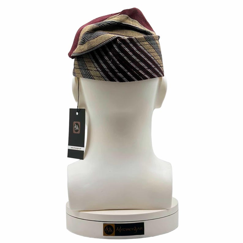 May include: A multi-colored, patterned hat with a tag. The hat is displayed on a white mannequin head. The hat features a combination of burgundy, tan, and black stripes and patterns. The tag is black with a logo and text.