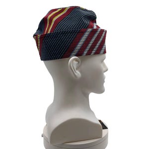 May include: A patterned hat with a rectangular brim. The hat features stripes of red, navy blue, yellow, and white. The hat is displayed on a white mannequin head.