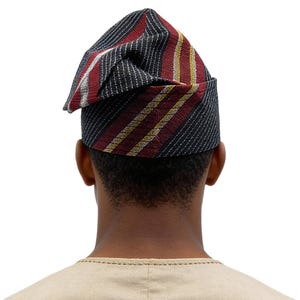 May include: A traditional African hat, seen from behind. The hat has a striped pattern in black, red, white, and yellow. The person is wearing a beige shirt. The hat is a cultural garment.