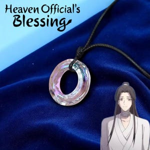 以下が含まれることがあります： ピンクと紫の虹色の色合いを持つ透明な円形ペンダントが、黒いコードから吊り下げられています。ペンダントは濃い青色の背景に配置されています。上部のテキストは「Heaven Official's Blessing」と書かれています。