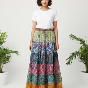 May include: A woman wearing a white t-shirt and a long, tiered skirt with a colorful paisley patchwork design. The skirt features a drawstring waist and a variety of colors including blue, pink, yellow, and green. The skirt is floor-length.