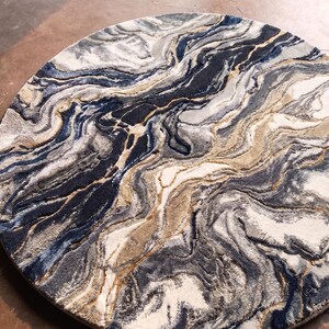 May include: A round rug with a marbled design. The rug features swirling patterns in shades of navy blue, gray, beige, and white. The design resembles the look of natural stone or flowing water. The rug is a decorative floor covering.