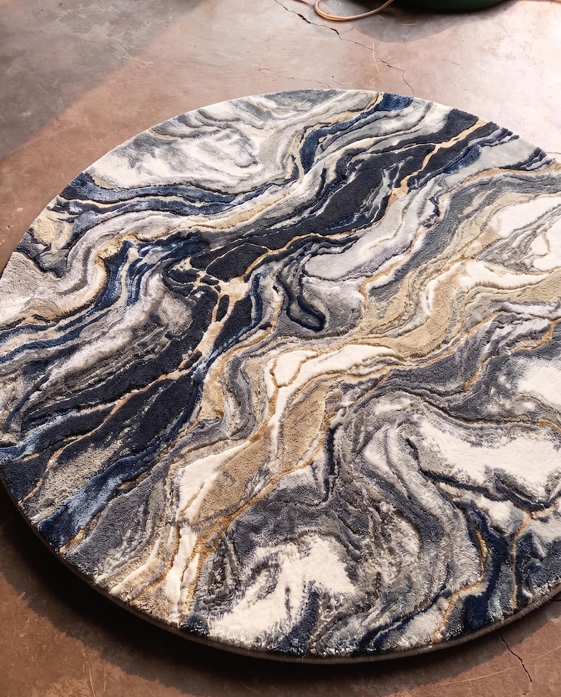 May include: A round rug with a marbled design. The rug features swirling patterns in shades of navy blue, gray, beige, and white. The design resembles the look of natural stone or flowing water. The rug is a decorative floor covering.