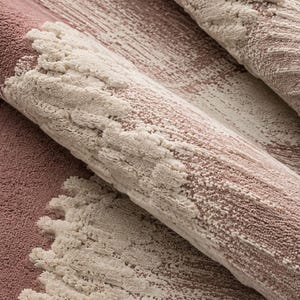 Op de afbeelding: Close-up van een opgerold tapijt met een textuurontwerp. Het tapijt heeft een mix van stoffige roze en gebroken witte kleuren. Het ontwerp bevat penseelstreekachtige patronen en verhoogde, getextureerde elementen, waardoor een gelaagd effect ontstaat.