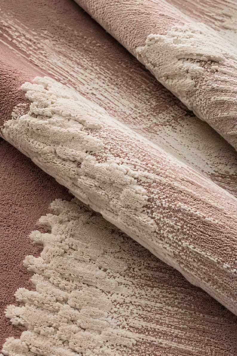Op de afbeelding: Close-up van een opgerold tapijt met een textuurontwerp. Het tapijt heeft een mix van stoffige roze en gebroken witte kleuren. Het ontwerp bevat penseelstreekachtige patronen en verhoogde, getextureerde elementen, waardoor een gelaagd effect ontstaat.
