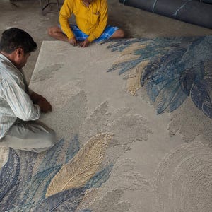Op de afbeelding: Een groot, rechthoekig tapijt met een veermotief in tinten blauw, beige en grijs. Twee mensen werken aan het tapijt, dat met de hand gemaakt lijkt te zijn. Rollen stof zijn zichtbaar op de achtergrond.