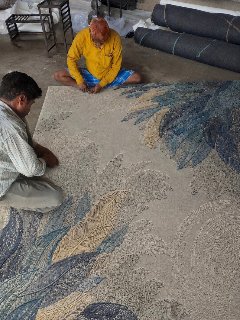 Op de afbeelding: Een groot, rechthoekig tapijt met een veermotief in tinten blauw, beige en grijs. Twee mensen werken aan het tapijt, dat met de hand gemaakt lijkt te zijn. Rollen stof zijn zichtbaar op de achtergrond.