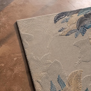 Op de afbeelding: Beige rechthoekig tapijt met een verhoogd veermotief in tinten blauw en bruin. Het tapijt heeft een getextureerd oppervlak en een gedetailleerd patroon, geschikt voor interieurdecoratie.