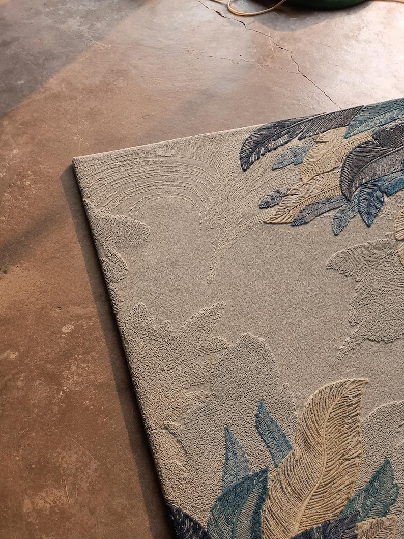 Op de afbeelding: Beige rechthoekig tapijt met een verhoogd veermotief in tinten blauw en bruin. Het tapijt heeft een getextureerd oppervlak en een gedetailleerd patroon, geschikt voor interieurdecoratie.