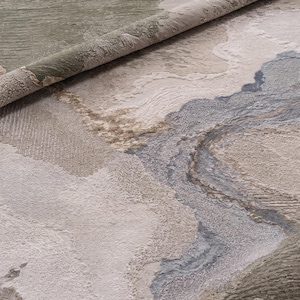 May include: A textured area rug featuring an abstract pattern with organic shapes in shades of sage green, cream, beige, and slate gray. The surface displays varying pile heights and directional weaves that create a layered, topographical appearance.