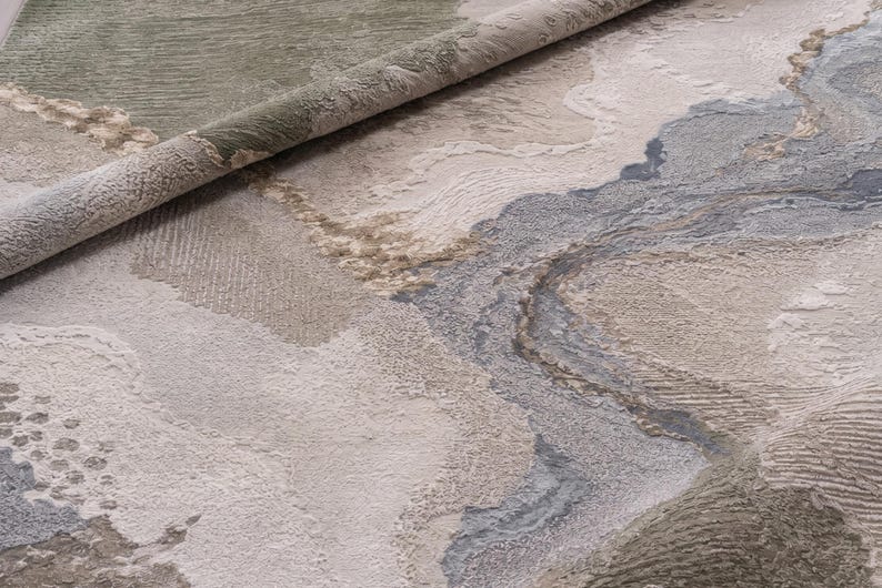 May include: A textured area rug featuring an abstract pattern with organic shapes in shades of sage green, cream, beige, and slate gray. The surface displays varying pile heights and directional weaves that create a layered, topographical appearance.
