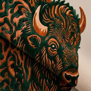 Peut inclure: Un tapis de bison vert et orange d&eacute;taill&eacute;. Le tapis pr&eacute;sente un motif de t&ecirc;te de bison stylis&eacute; avec des motifs et des textures complexes. Les cornes et les d&eacute;tails du visage du bison sont mis en &eacute;vidence en orange sur un fond vert.
