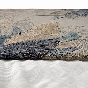 Op de afbeelding: Een close-up van een getextureerd tapijt met een veermotief. Het tapijt heeft grote, gedetailleerde veren in tinten blauw, beige en grijs. De achtergrond heeft een subtiel, getextureerd patroon. Het tapijt is waarschijnlijk gemaakt van een zacht, duurzaam materiaal.