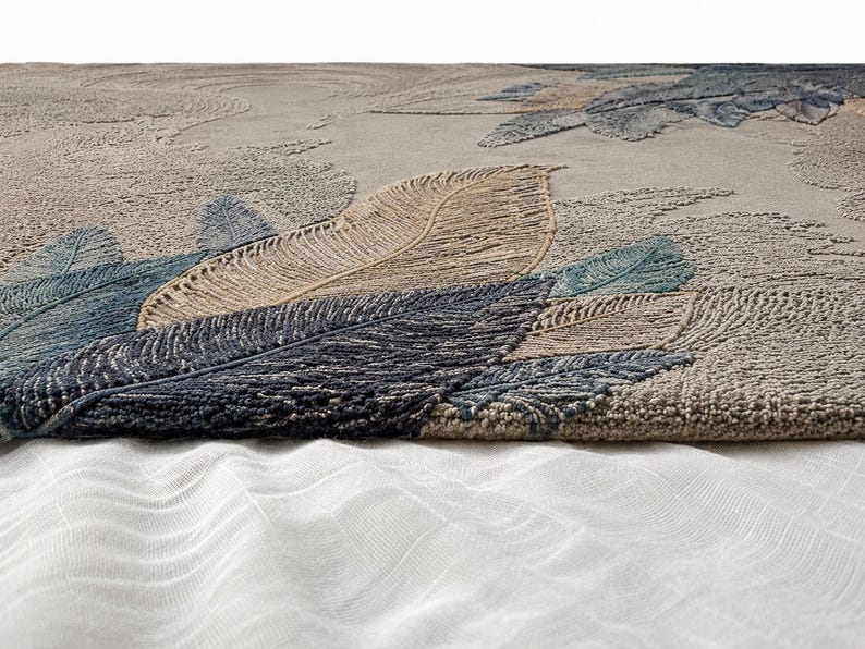 Op de afbeelding: Een close-up van een getextureerd tapijt met een veermotief. Het tapijt heeft grote, gedetailleerde veren in tinten blauw, beige en grijs. De achtergrond heeft een subtiel, getextureerd patroon. Het tapijt is waarschijnlijk gemaakt van een zacht, duurzaam materiaal.