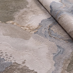 May include: A textured area rug featuring an abstract pattern with swirling organic shapes in shades of beige, taupe, cream, and slate blue. The surface displays varying pile heights and carved details that create a multi-dimensional appearance.