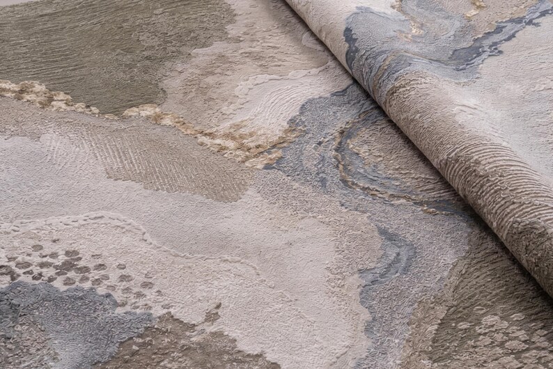 May include: A textured area rug featuring an abstract pattern with swirling organic shapes in shades of beige, taupe, cream, and slate blue. The surface displays varying pile heights and carved details that create a multi-dimensional appearance.