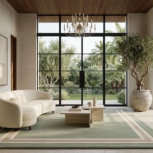 May include: A living room featuring a curved cream-colored sofa, a two-tiered travertine coffee table, and a large rectangular area rug in sage green with beige borders. A tall olive tree in a textured stone pot stands near floor-to-ceiling windows.