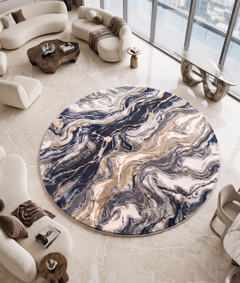 May include: A round area rug with a marbled design in shades of blue, gray, and beige. The rug is placed on a light-colored marble floor, surrounded by modern furniture. The rug's pattern resembles flowing water or stone.