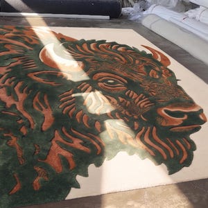 Peut inclure: Un tapis rectangulaire avec une illustration d&eacute;taill&eacute;e d'une t&ecirc;te de bison. Le design incorpore des nuances de marron, de vert et de cr&egrave;me. La t&ecirc;te du bison est bien en &eacute;vidence, avec des d&eacute;tails complexes dans la fourrure et les cornes. Le tapis est probablement fait d'une mati&egrave;re douce.