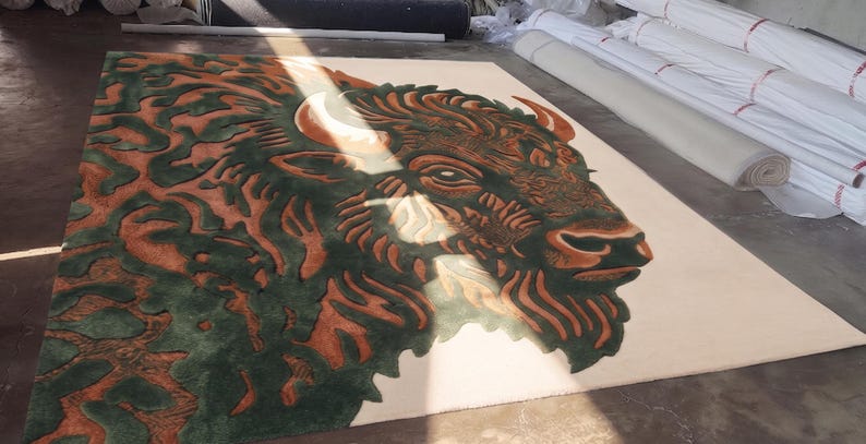 Peut inclure: Un tapis rectangulaire avec une illustration d&eacute;taill&eacute;e d'une t&ecirc;te de bison. Le design incorpore des nuances de marron, de vert et de cr&egrave;me. La t&ecirc;te du bison est bien en &eacute;vidence, avec des d&eacute;tails complexes dans la fourrure et les cornes. Le tapis est probablement fait d'une mati&egrave;re douce.