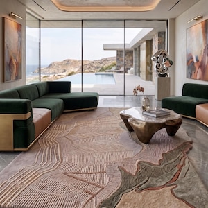 May include: A large rectangular area rug featuring a textured, multi-layered design with abstract topographical patterns in shades of beige, tan, muted green, and soft pink, covering a polished gray marble floor in a spacious living room setting.